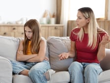 Angry teenager sits on the couch turned away from her mother who is trying to talk to her