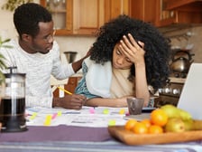 A young man comforts a stressed young woman reviewing papers at a kitchen table.