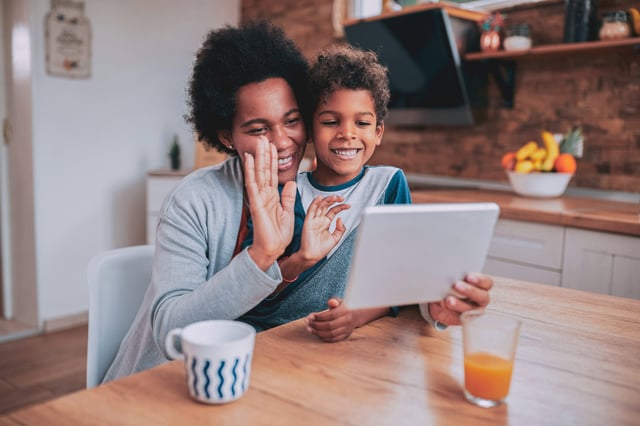 An adult woman and a young boy are smiling and waving while looking at a tablet together in a bright kitchen. They appear happy and engaged in a video call or digital activity.