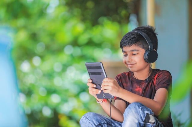 A young boy wearing black headphones is smiling while looking at a digital tablet or large smartphone. He is seated outdoors, with a blurred green leafy background, appearing calm and engaged with his device.