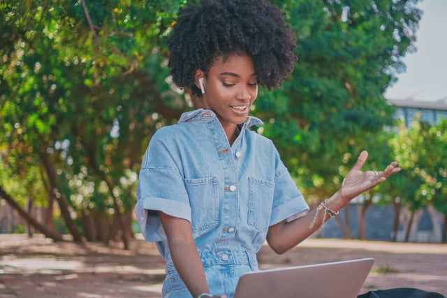 Young adult female with dark skin and a jean jacket at her laptop outside