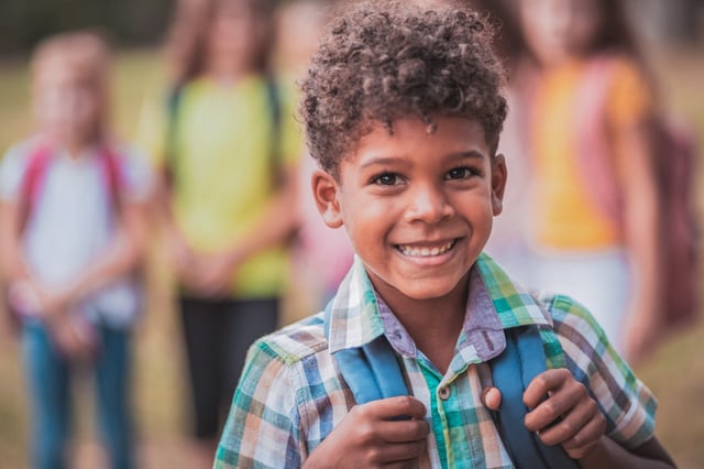 A young child, wearing a plaid shirt and a blue backpack, smiles broadly at the camera. Other children are visible, blurred, in the background outdoors, suggesting a school or play environment.