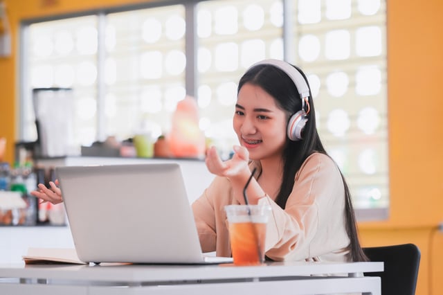 A smiling teen wearing headphones talks animatedly during a video call on a laptop in a bright, modern indoor setting. A drink sits on the table beside the laptop.