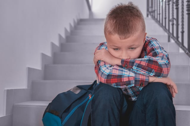 A young boy sits alone on a set of indoor stairs, appearing sad and withdrawn with his head down and arms resting on his knees. A backpack is visible next to him on the stairs.