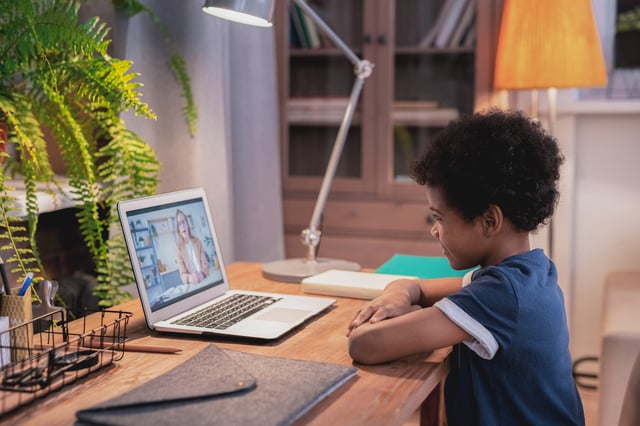 A young child, appearing about 5-8 years old, is seated at a wooden desk, looking intently at a laptop screen. An adult therapist is visible on the video call, suggesting an online therapy session in a calm, indoor setting.