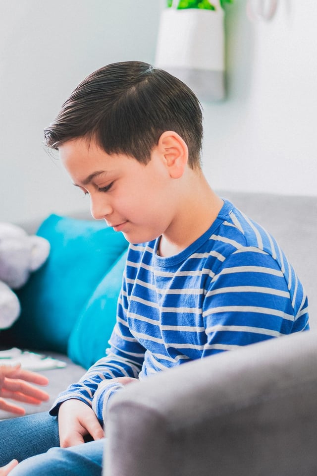 A young boy wearing a blue and white striped shirt sits on a sofa, looking down intently at something in front of him. An adult's hands are gently placed on his leg, suggesting a supportive and comforting interaction in a relaxed indoor setting.