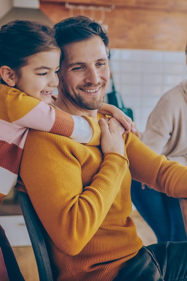 A happy family is gathered in a bright home setting. A young girl hugs an adult man who is holding a phone, while another young girl colors at the table, and an adult woman smiles in the background.