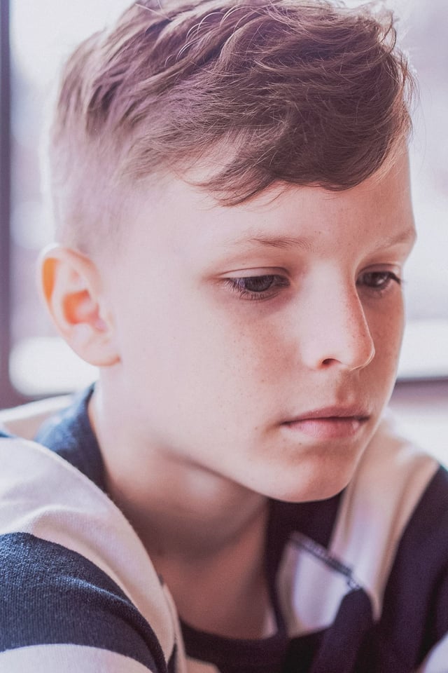 A close-up portrait of a young boy with short hair and freckles, looking down with a contemplative expression in a softly lit indoor setting. He appears to be deep in thought.