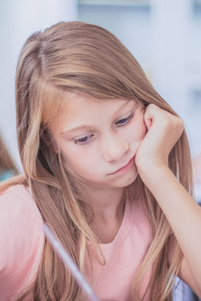A young girl with long, light brown hair sits at a desk, concentrating while writing or drawing and resting her chin in her hand. She appears focused and thoughtful, likely in a classroom or study environment.