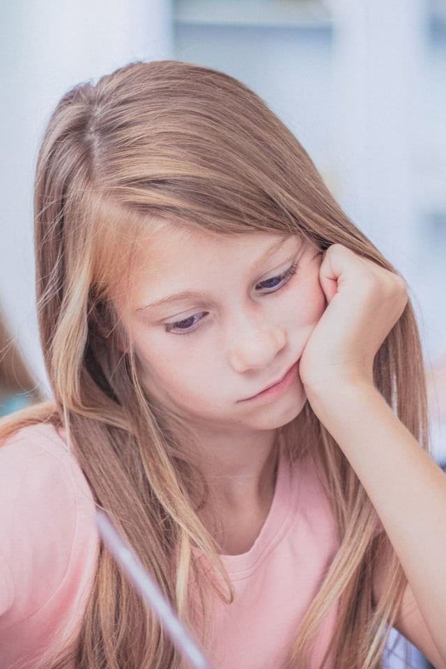A young girl with long, light brown hair sits at a desk, concentrating while writing or drawing and resting her chin in her hand. She appears focused and thoughtful, likely in a classroom or study environment.