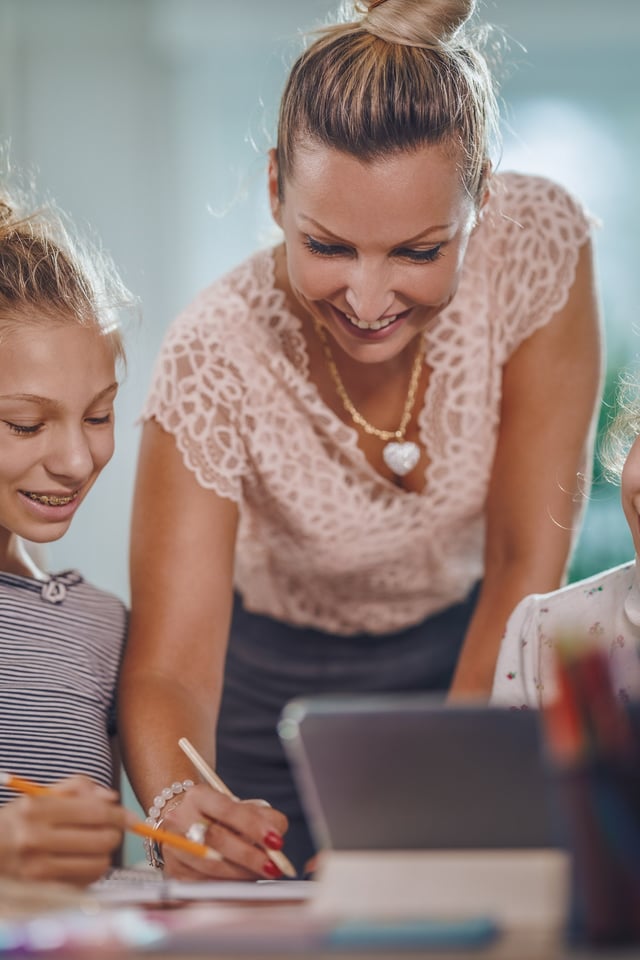 An adult woman smiles as she guides a pre-teen and a younger child in a learning activity at a table. They are writing and looking at a tablet in a bright home setting.