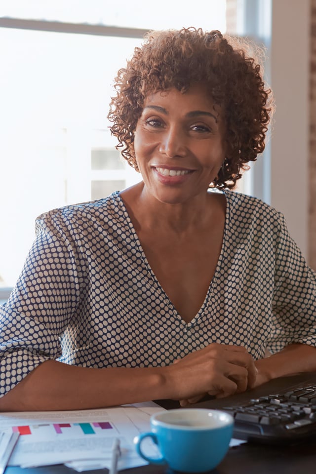 A smiling adult woman with curly hair sits at a desk, looking directly at the camera in a brightly lit office setting. A computer monitor, documents, and a coffee cup are visible on the desk, with a large window and brick wall in the background.