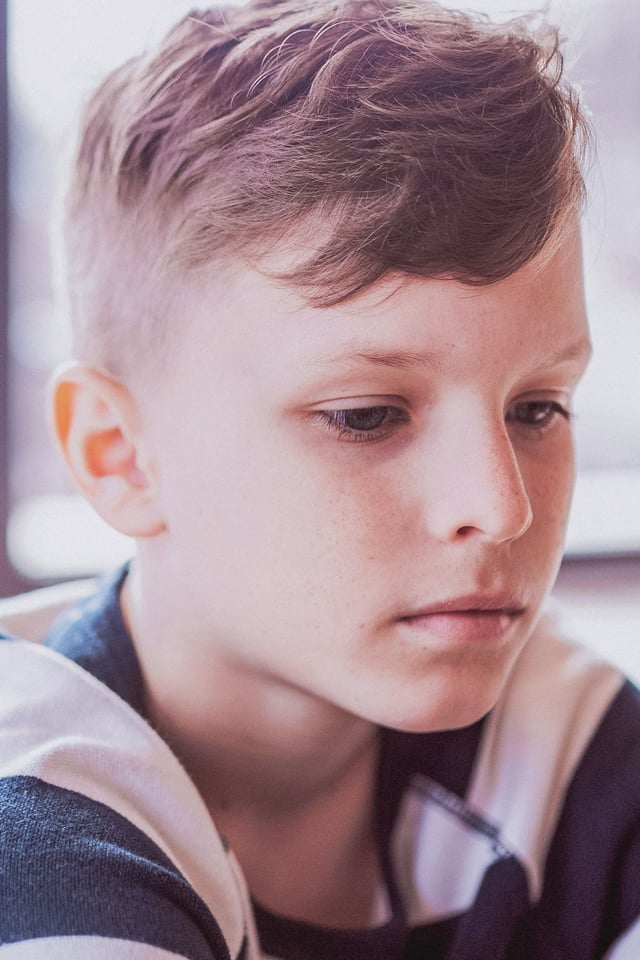 A close-up portrait of a young boy, appearing to be a pre-teen, looking down with a pensive and thoughtful expression. He is wearing a striped shirt indoors, with soft lighting on his face.