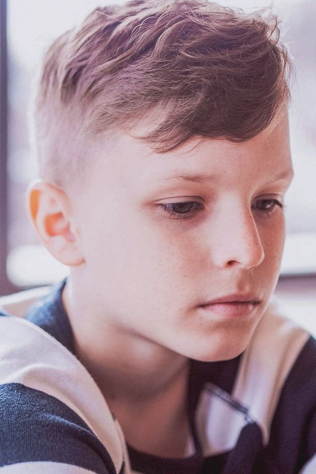 A close-up portrait of a young boy with short hair and freckles, looking down with a contemplative expression in a softly lit indoor setting. He appears to be deep in thought.