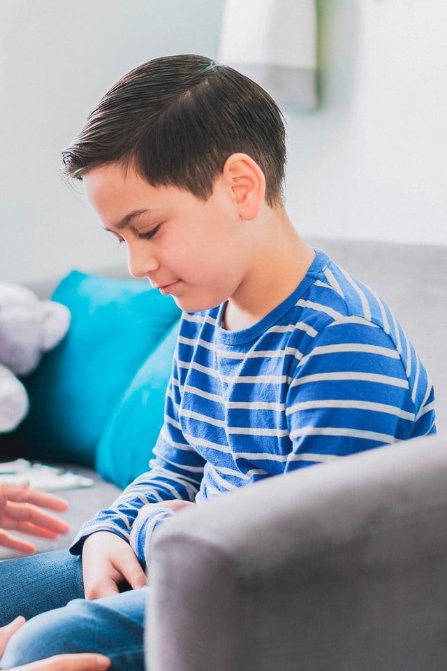 A young boy wearing a blue and white striped shirt sits on a sofa, looking down intently at something in front of him. An adult's hands are gently placed on his leg, suggesting a supportive and comforting interaction in a relaxed indoor setting.
