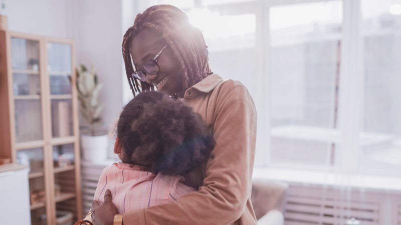 An adult with dark braids and eyeglasses is embracing a child in a brightly lit indoor space. The adult is smiling warmly, conveying a sense of comfort and affection.