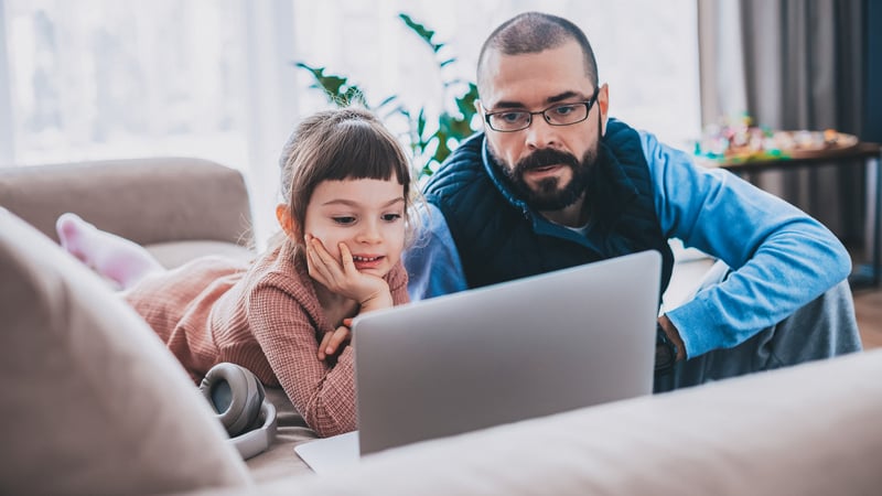 A young girl and an adult man are engaged, looking at a laptop screen together in a cozy living room. The girl lies on a sofa with headphones nearby, while the man sits attentively beside her.