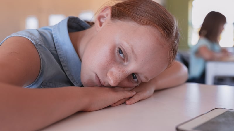 A young girl with reddish-brown hair rests her head on her arms on a table, looking directly at the viewer with a subdued and thoughtful expression. She is in an indoor setting, possibly a classroom, with a blurred figure seated at a desk in the background.