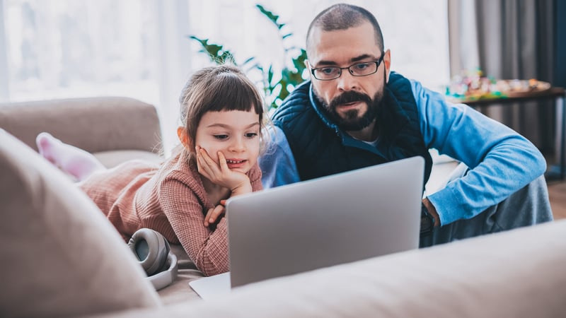 A young girl and an adult man are engaged, looking at a laptop screen together in a cozy living room. The girl lies on a sofa with headphones nearby, while the man sits attentively beside her.