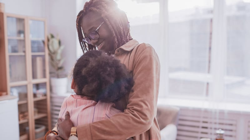 An adult with dark braids and eyeglasses is embracing a child in a brightly lit indoor space. The adult is smiling warmly, conveying a sense of comfort and affection.