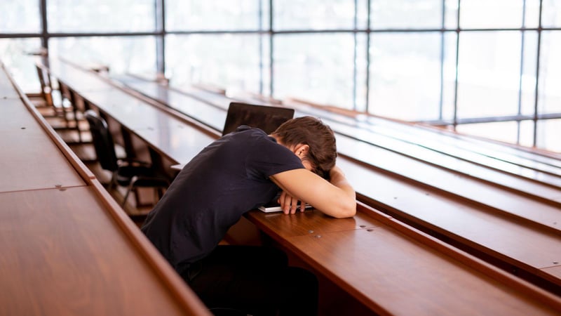 A teen sits alone in an empty lecture hall, resting their head face-down on their desk in exhaustion or distress.