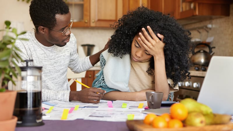 A young man comforts a stressed young woman reviewing papers at a kitchen table.