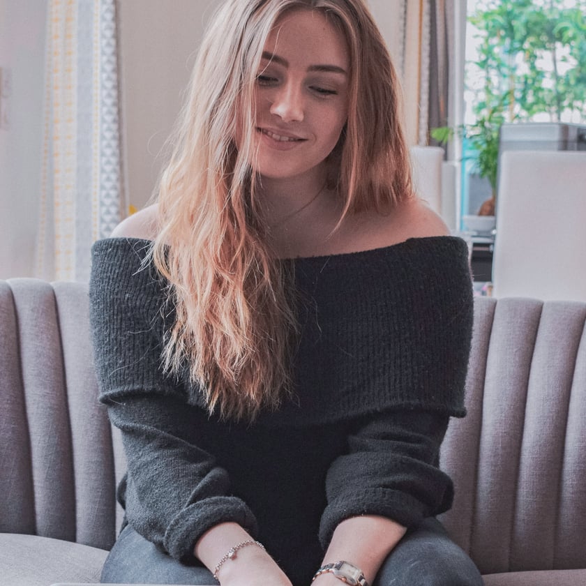 A young woman or teen sits comfortably on a grey couch, looking down at a laptop with a gentle smile, indicating an engaged online interaction in a bright indoor setting.