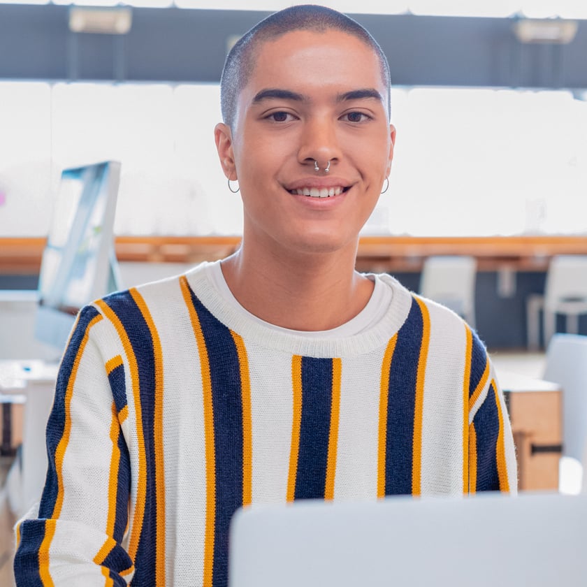 A smiling young adult with a shaved head and nose piercing looks directly at the camera while sitting at a desk with a laptop in a bright, modern indoor setting. They appear friendly and engaged.