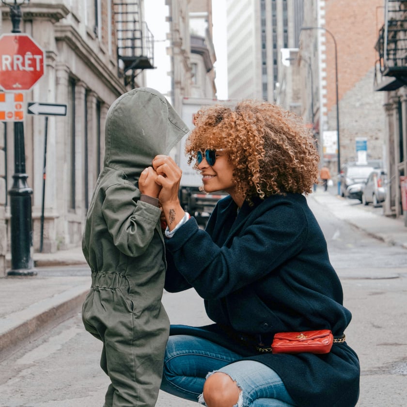 An adult with curly hair and sunglasses crouches down to hold the hands of a young child in a hooded jumpsuit on an urban street. The adult is smiling warmly at the child, conveying a sense of connection and care.