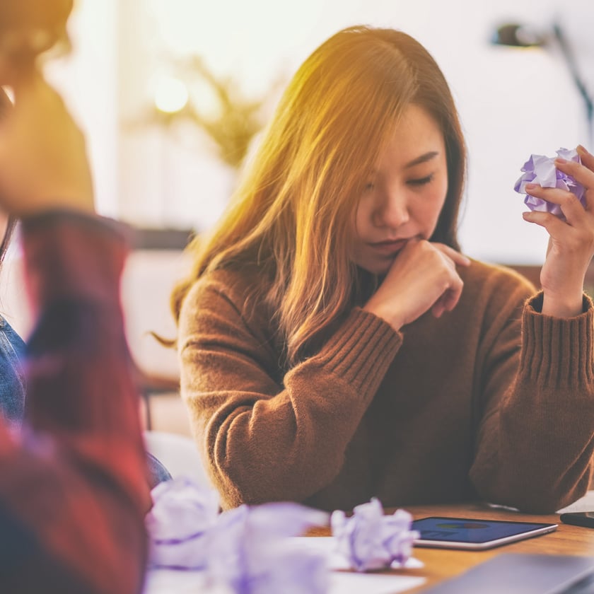 An adult woman looks down contemplatively, holding a crumpled piece of paper, while another adult and a teen in the background also appear somber or distressed around a table indoors. The scene conveys a mood of frustration or deep thought.