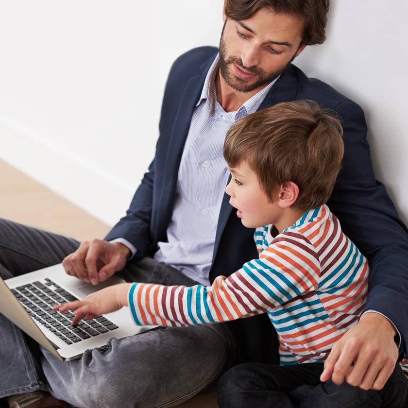 An adult man and a young boy are sitting on a light wood floor, leaning against a white wall, both looking at a laptop. The boy is pointing at the laptop while the man watches him attentively.