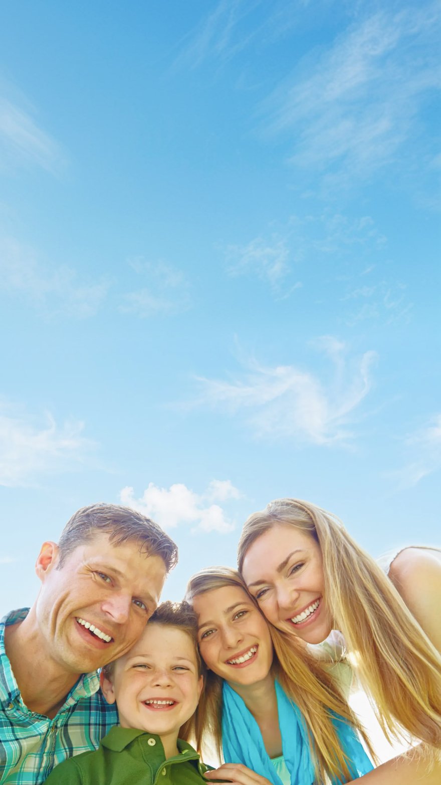 A cheerful family of four — mother, father, a young boy, and their older daughter — huddled close and smiling up at the camera on a bright, sunny day.