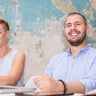 Two smiling adults are seated together in an indoor setting with a distressed blue and white wall behind them. A woman with short orange hair is seated at a laptop, while a man with a beard sits next to her, looking happy and engaged.