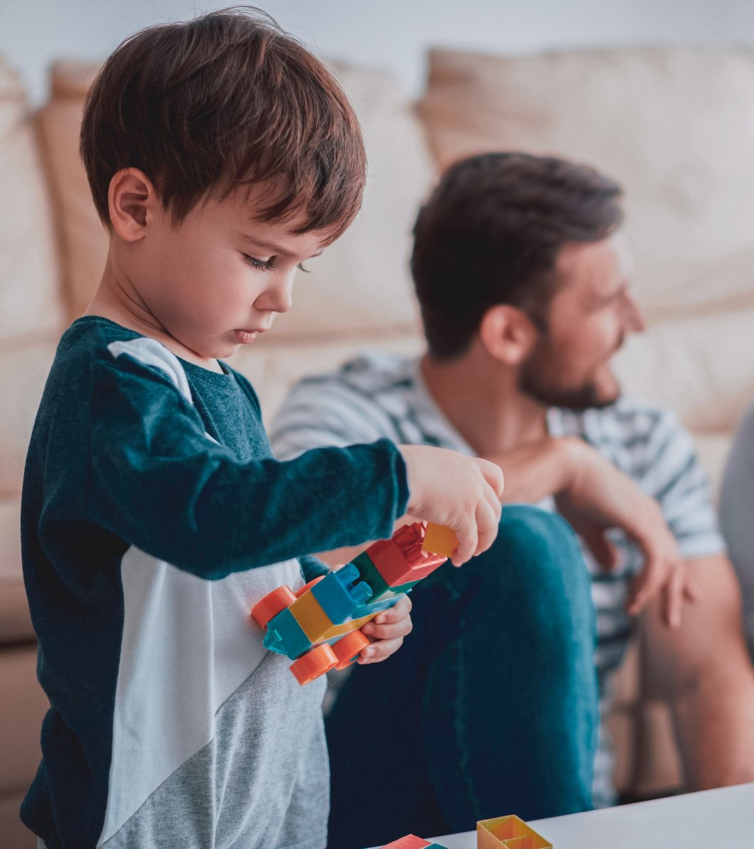 A young child is focused on playing with colorful building blocks at a white table. In the softly blurred background, an adult male and female sit on a sofa, appearing to be in a calm conversation in an indoor setting.