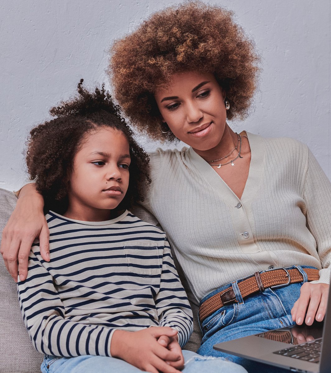 An adult woman with curly hair has her arm around a child with curly hair as they both sit on a sofa. They are attentively looking at a laptop screen, with the child's expression appearing pensive while the adult looks supportive.