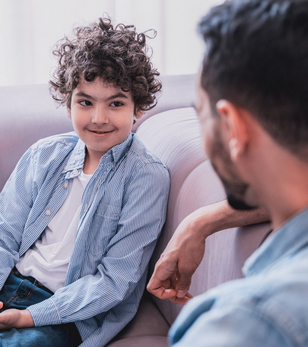 A young boy with curly hair sits on a light-colored sofa, looking up and smiling subtly at an adult. The adult, seen from behind, is leaning towards the boy, engaged in a calm conversation in an indoor setting.