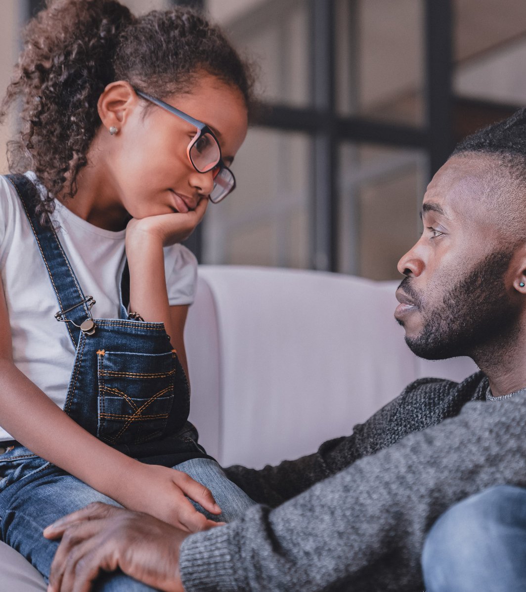A young girl with curly hair and glasses sits on an adult man's lap on a couch, looking up at him with a pensive expression. The man, who has dreadlocks, looks back at her attentively, his arm around her in a comforting gesture, suggesting a serious conversation or moment of support.