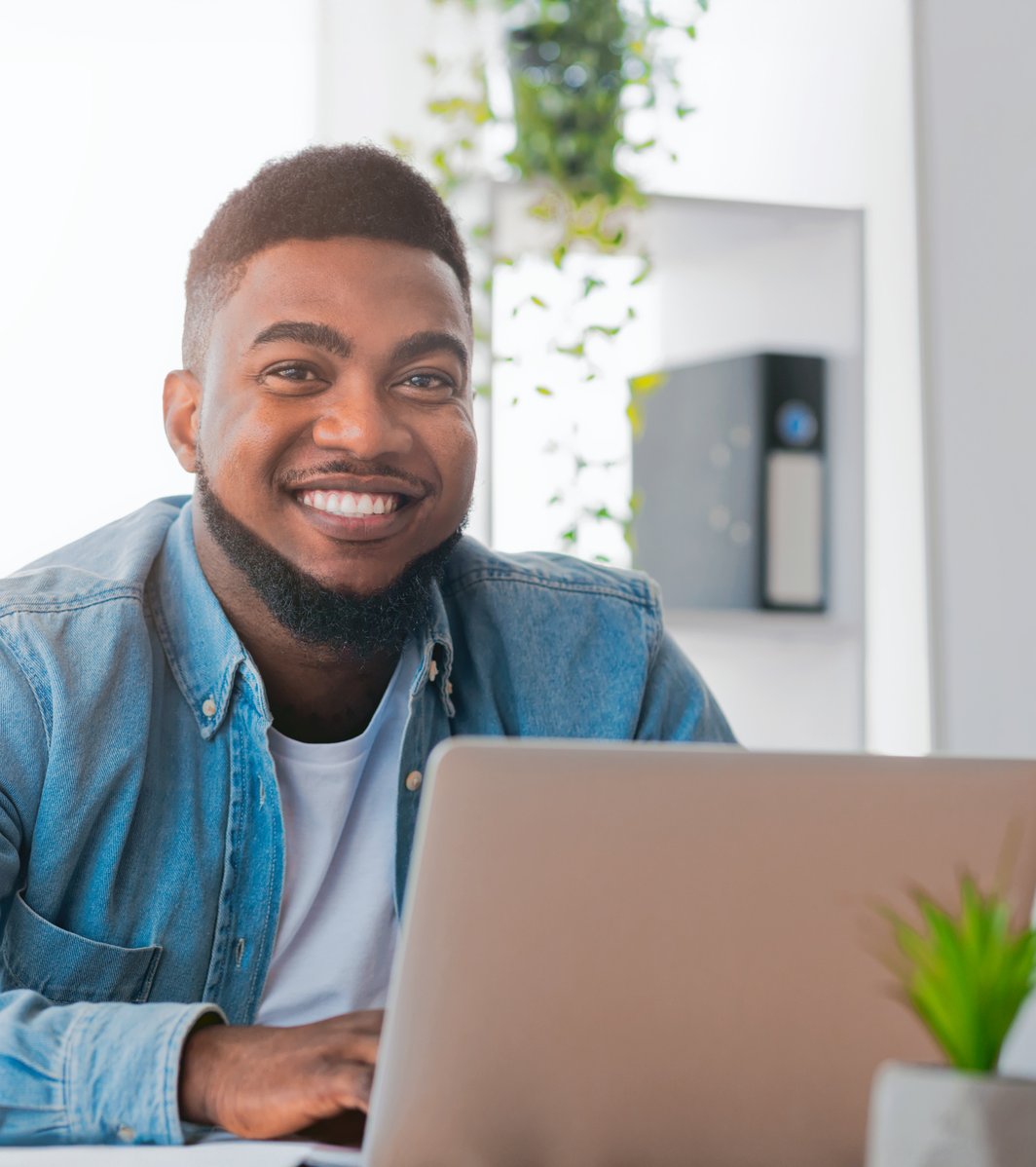 A smiling adult man sits at a desk with a laptop in a bright, modern office setting. He is looking directly at the camera with a friendly and approachable expression.