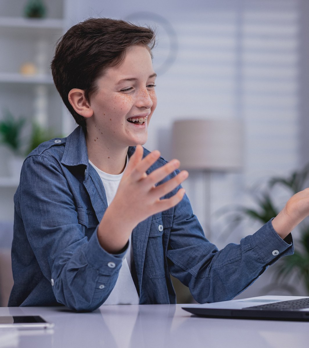 A happy teenager with braces is actively gesturing with both hands while looking at a laptop. They are seated at a desk in a bright indoor setting, seemingly engaged in a virtual conversation.