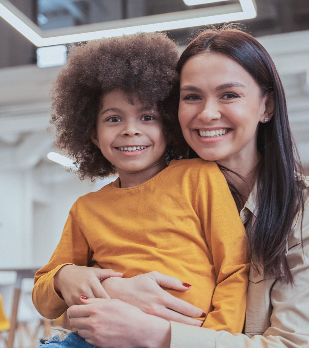 An adult woman warmly embraces a smiling young child, both looking directly at the camera. They are in a bright, modern indoor setting, conveying a mood of happiness and connection.