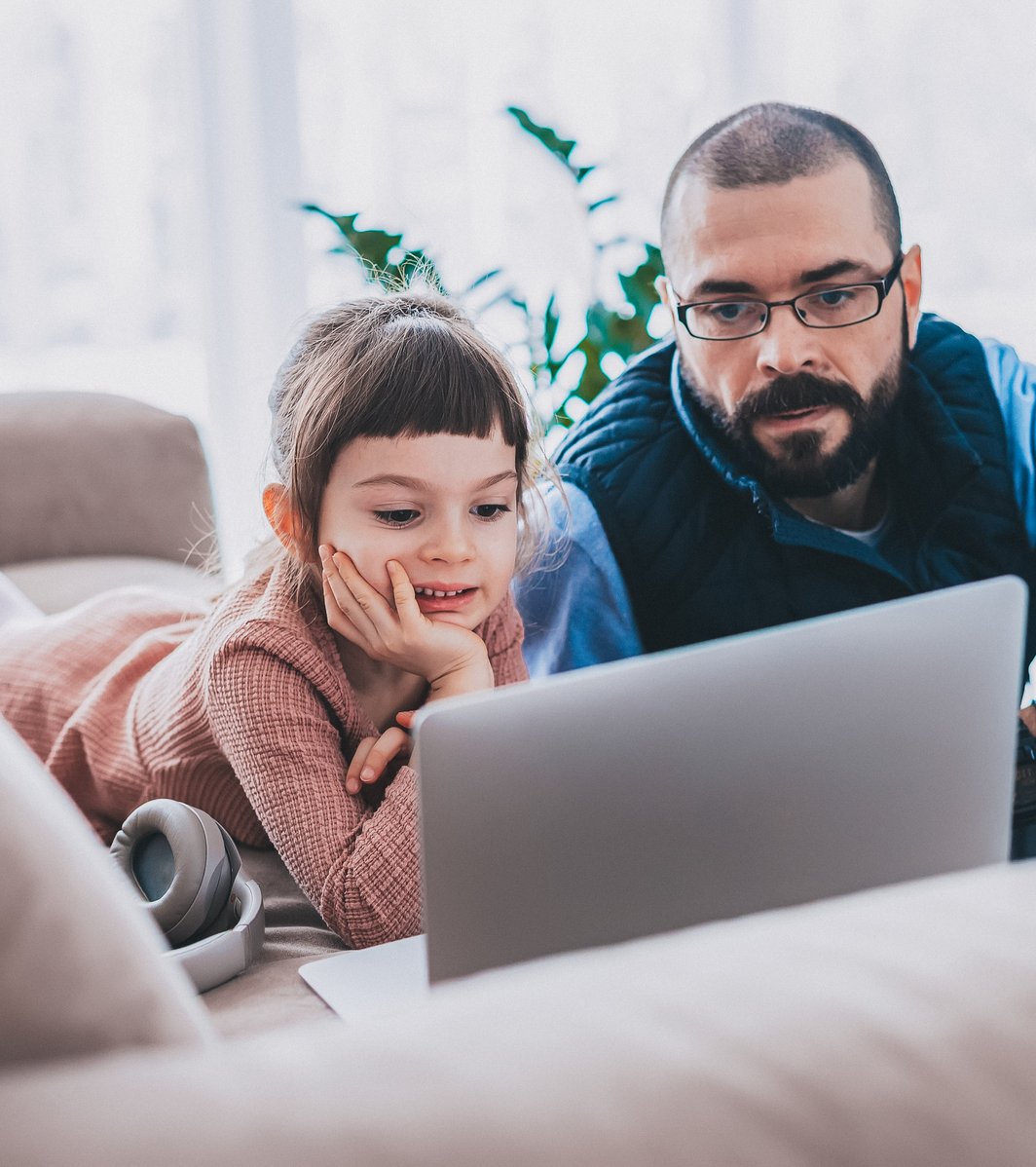 A young child and an adult man are intently focused on a laptop screen in a bright, cozy living room. The child lies comfortably on a sofa with headphones nearby, while the man sits on the floor beside her.