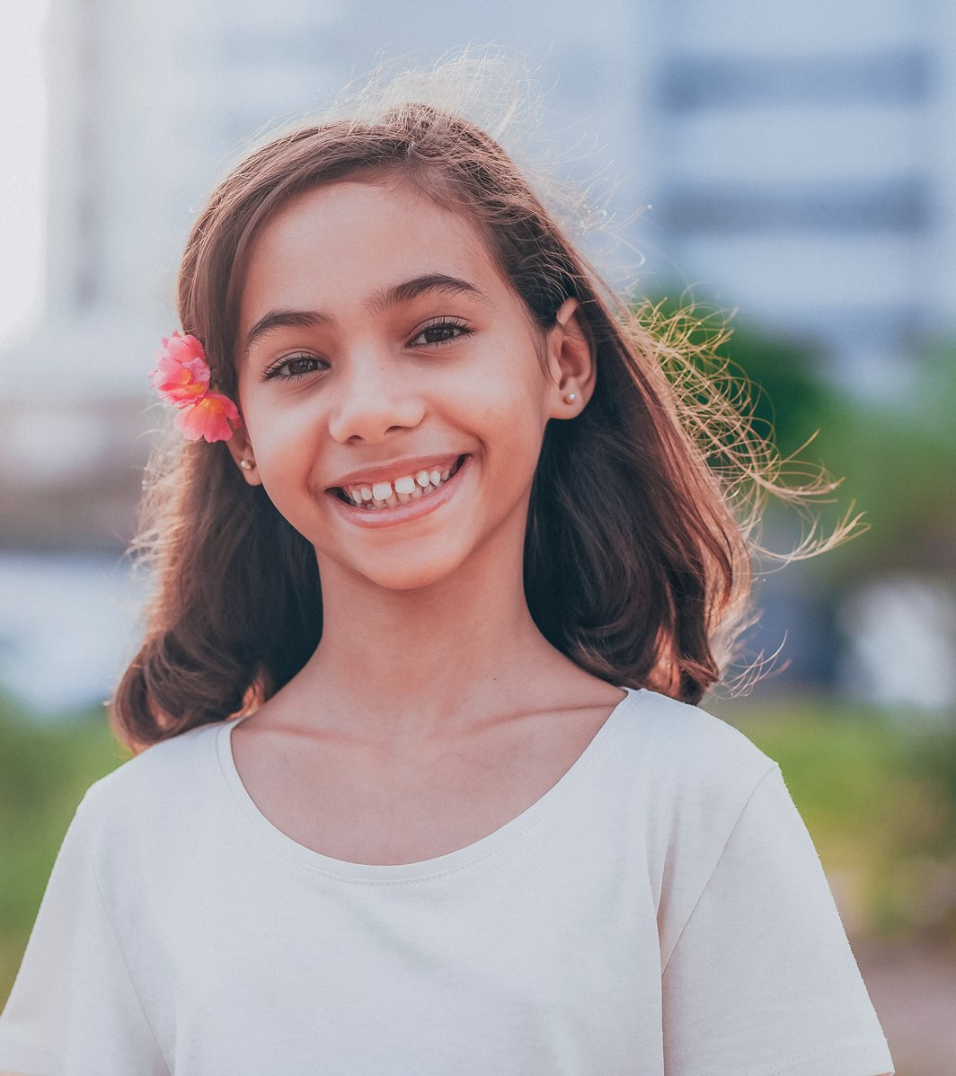 A young girl with long brown hair and a pink flower behind her ear smiles brightly. She is outdoors with a softly blurred green background.