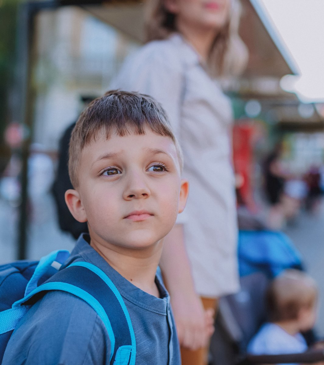 A young boy with a blue backpack stands outdoors in an urban setting, looking upwards with a thoughtful expression. An adult and a toddler in a stroller are blurred in the background, suggesting a family waiting.