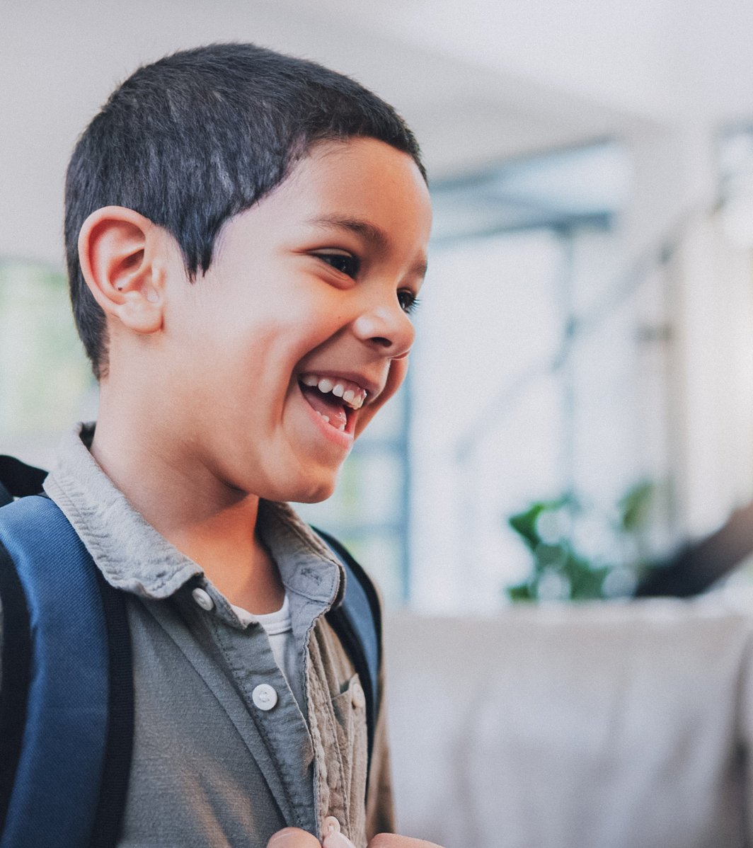 A young Hispanic/Latino boy, wearing a backpack, is laughing joyfully while looking up at an adult man in a bright, comfortable indoor setting. The scene conveys a warm, happy interaction.
