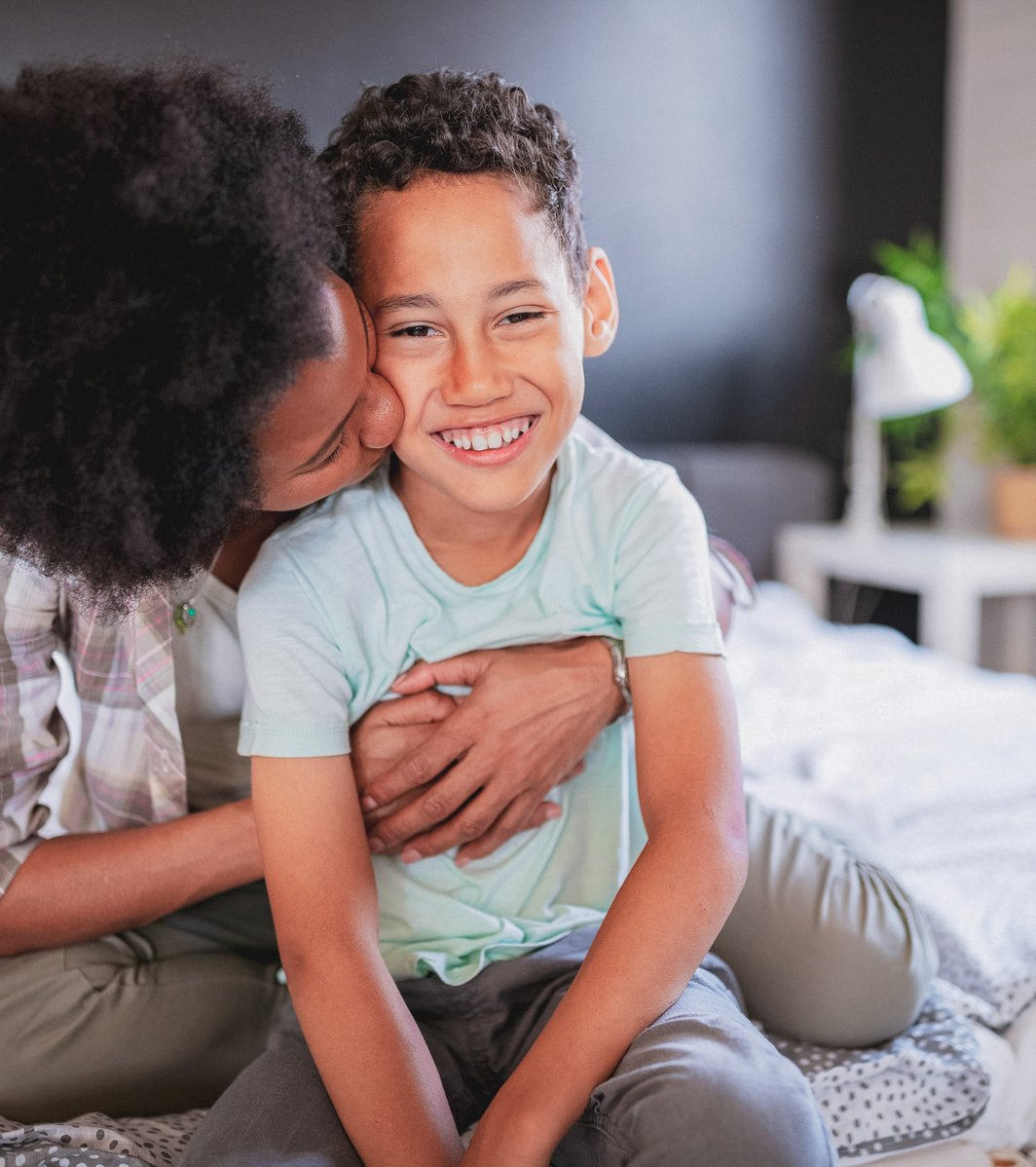 An adult is affectionately kissing a young child's cheek while sitting together on a bed. The child is smiling brightly at the camera, looking happy and secure.