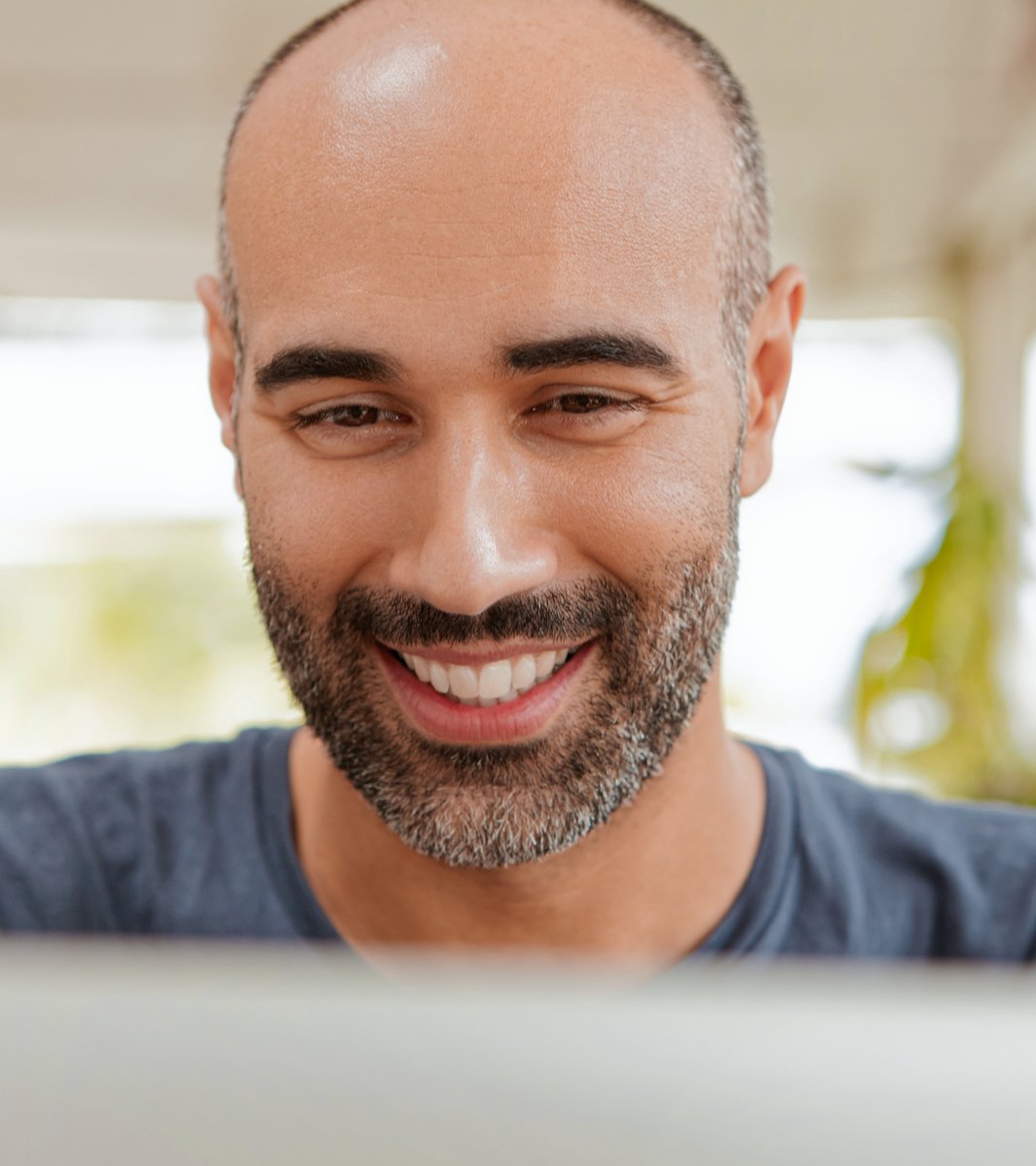 A smiling adult man with a beard looks down with a friendly and engaged expression. The background is softly blurred, suggesting a bright indoor or semi-outdoor setting.