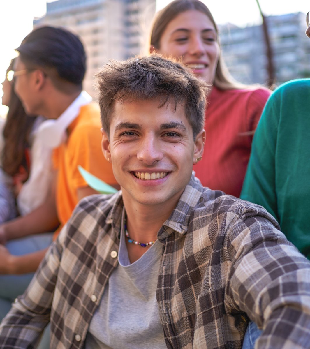 A smiling young man in a plaid shirt looks at the viewer, surrounded by a group of happy teens and young adults in an outdoor urban setting.