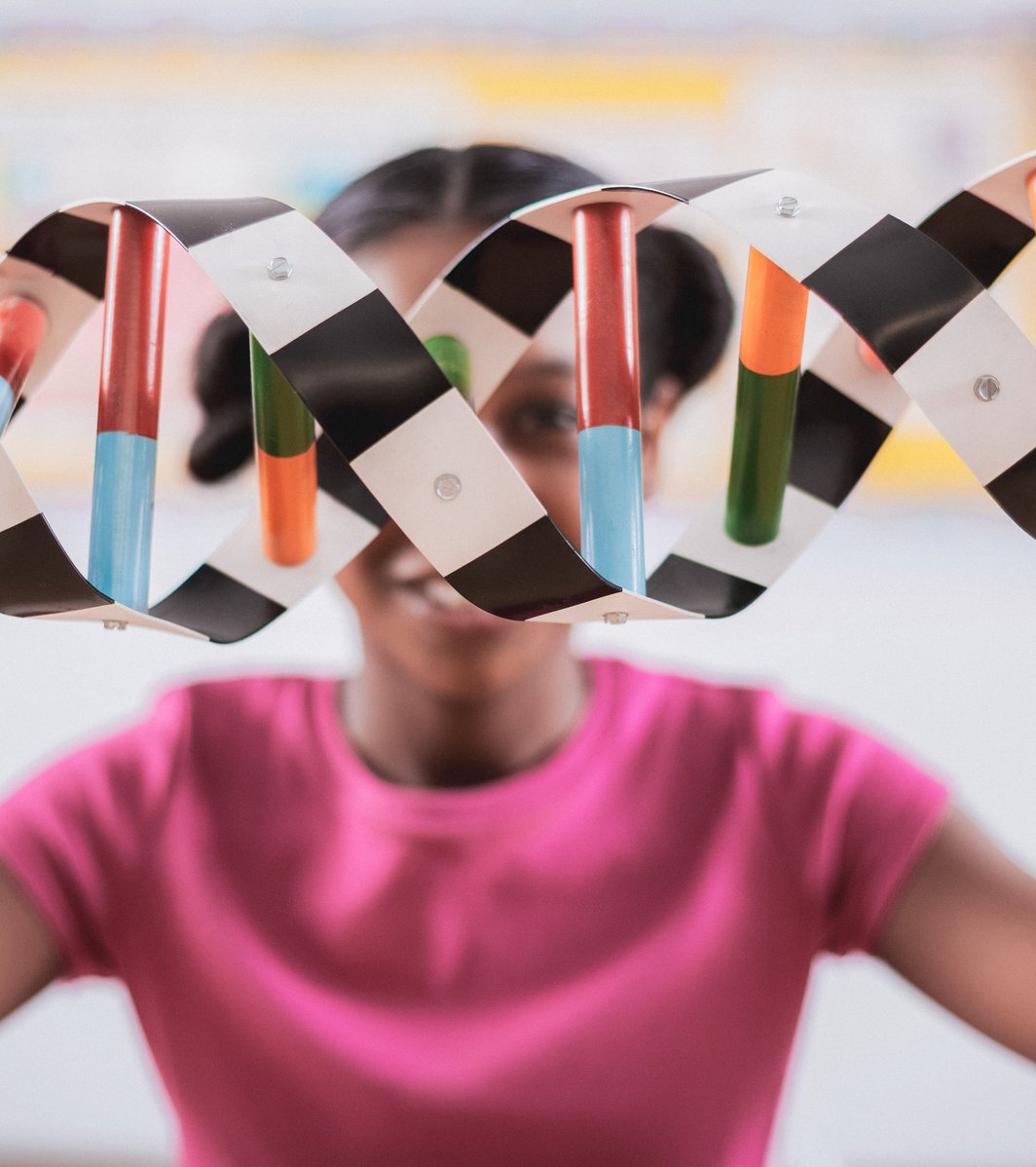 A young child holds up a large, colorful DNA model, appearing engaged and curious in a brightly lit, blurred classroom setting.