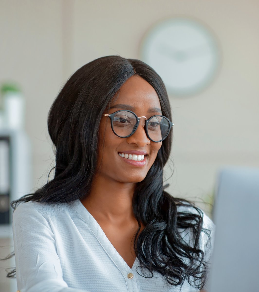 An adult woman with long dark hair and glasses is smiling warmly while looking attentively at a laptop screen. She appears to be in a professional office setting, conveying a friendly and engaged mood.