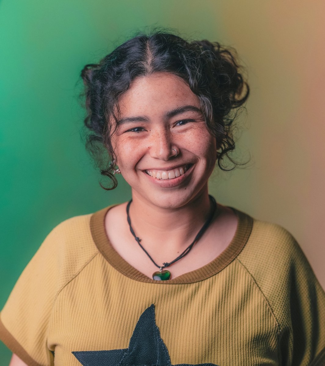 A smiling teen with curly hair and a nose piercing looks directly at the camera against a gradient green and orange background. They wear a ribbed shirt with a black star design, conveying a friendly and positive mood.